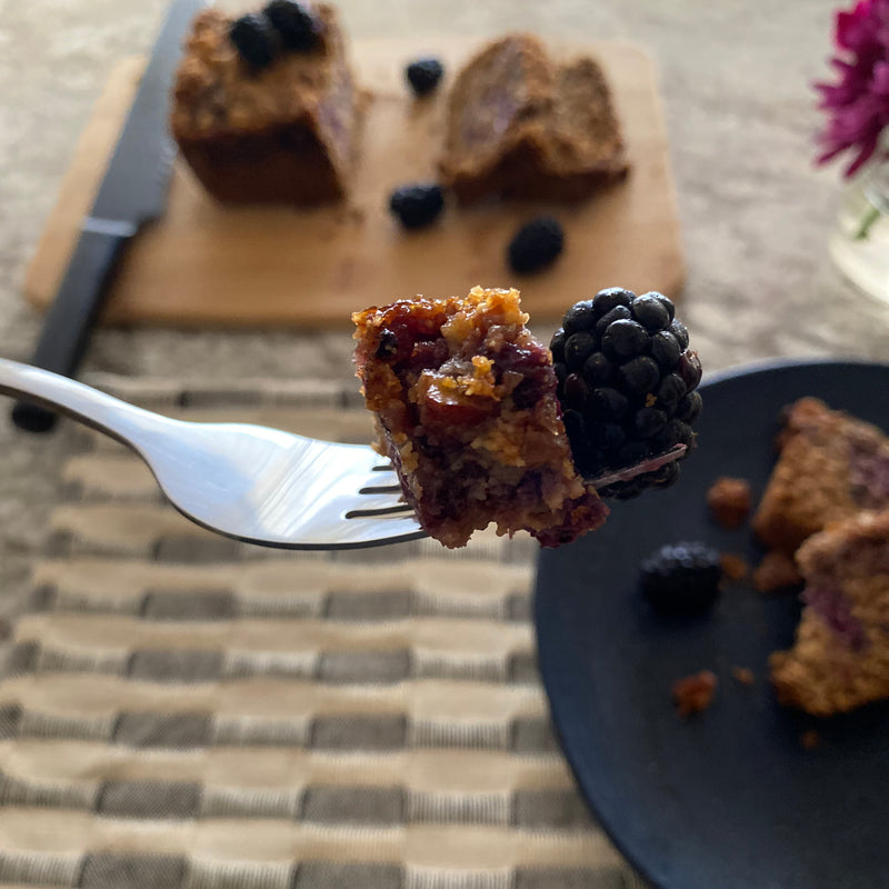 A moist and delicious bite of blackberry bread with fresh blackberries. Indulgent slices of the blackberry bread in the background are accompanied by more fresh blackberries and a vase of magenta flowers.